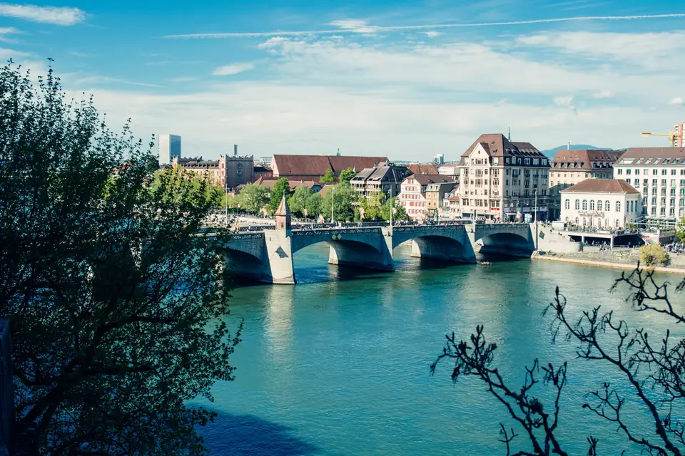 The Mittlere Brücke crossing the turquoise Rhine in Basel on a sunny day, with old riverside houses and a busy waterfront beyond
