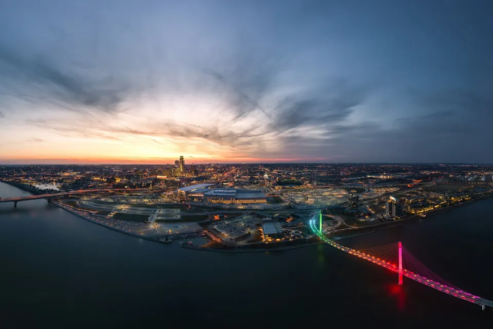 Omaha at dusk from above the Missouri River, with the downtown skyline, the riverfront arena district and a pedestrian bridge lit pink and green