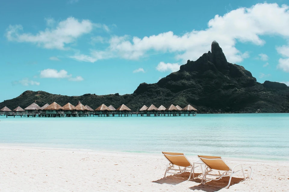 Two sun loungers on white sand facing thatched overwater bungalows and Mount Otemanu across the turquoise lagoon of Bora Bora