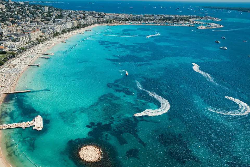Aerial view of the bay at Cannes along La Croisette, with turquoise water, jet-ski wakes, beach clubs and a marina at the far end of the beach