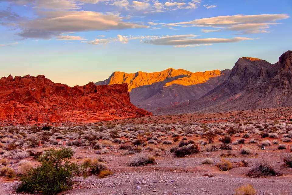 Red sandstone outcrops and rugged desert mountains outside Las Vegas, lit by the low sun under a blue sky with streaks of cloud