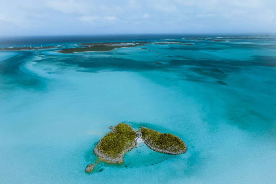 A small private cay with a lone house and jetty in shallow turquoise water, with more cays and sandbanks stretching to the horizon in the Bahamas