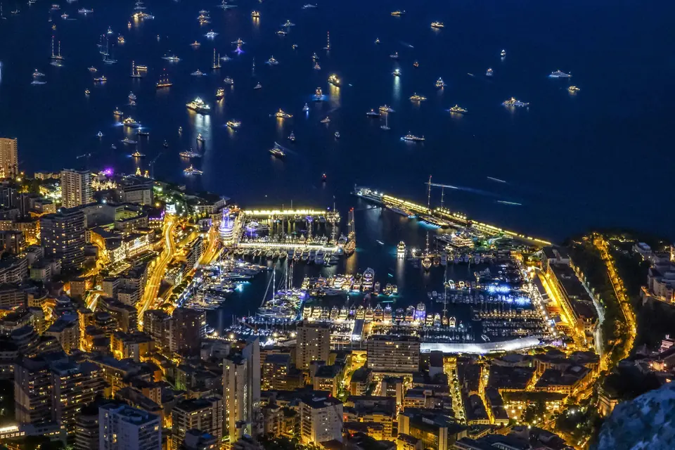 Monaco's Port Hercule at night, seen from high above the city, with the harbour lit gold and yachts anchored offshore