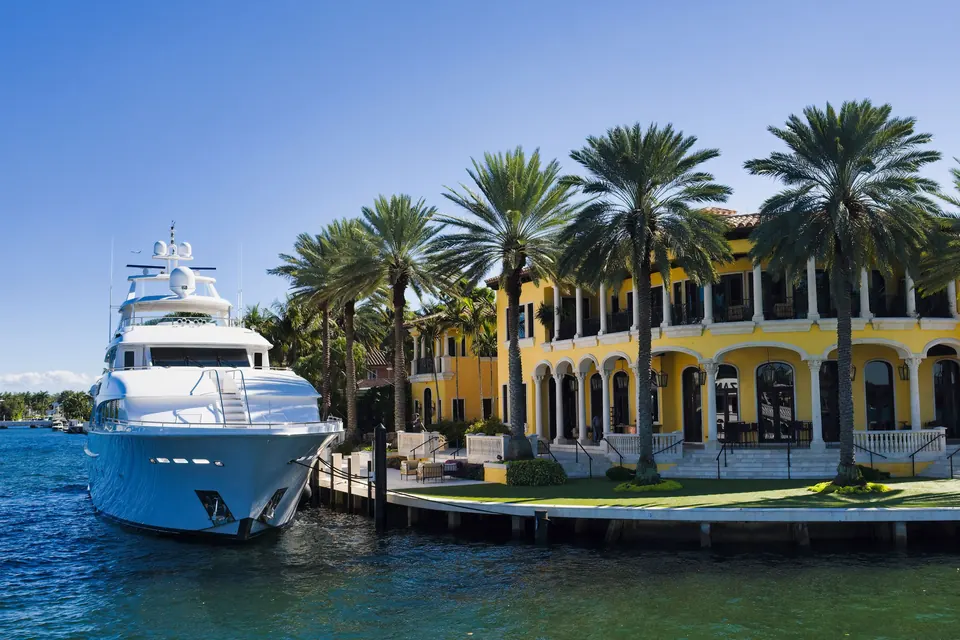 A white motor yacht moored beside a yellow waterfront mansion lined with palms in Fort Lauderdale