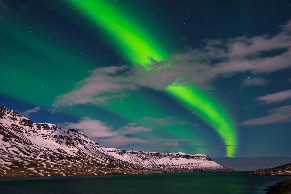 A bright green aurora arcing over snow-streaked mountains and a dark fjord at night in Iceland