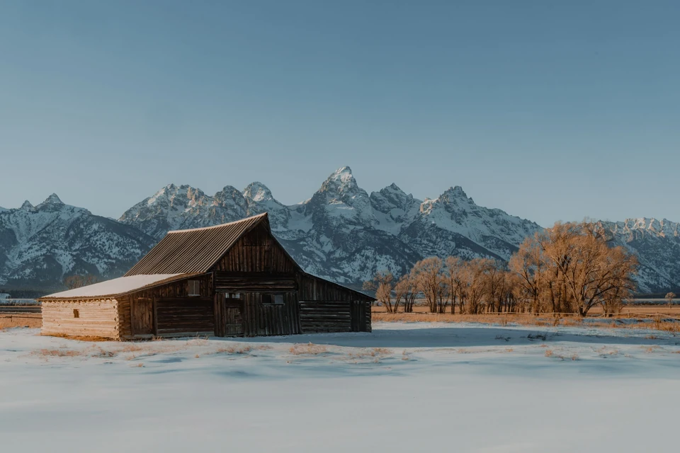 A historic wooden barn in deep snow beneath the snow-capped Teton Range in early-morning light, Jackson Hole, Wyoming