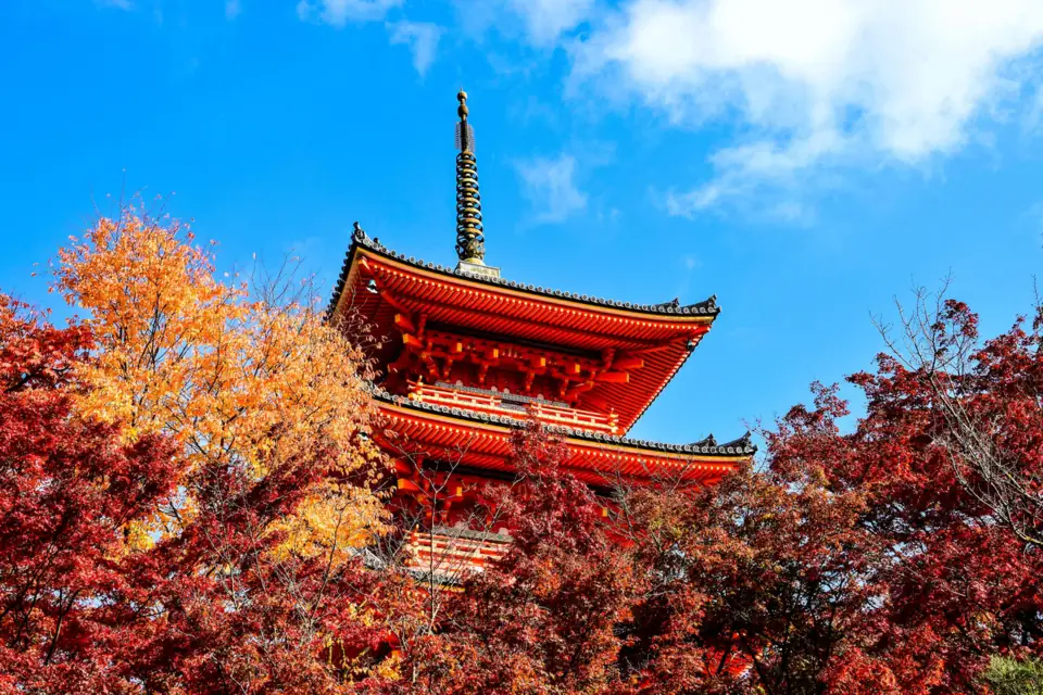 The vermilion pagoda of Kiyomizu-dera framed by red and gold autumn maples under a blue sky in Kyoto