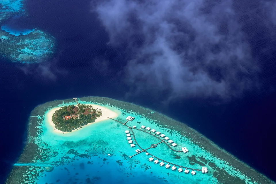 A small Maldives resort island ringed by white sand, with overwater villas on a turquoise reef lagoon and wisps of cloud over the deep-blue ocean