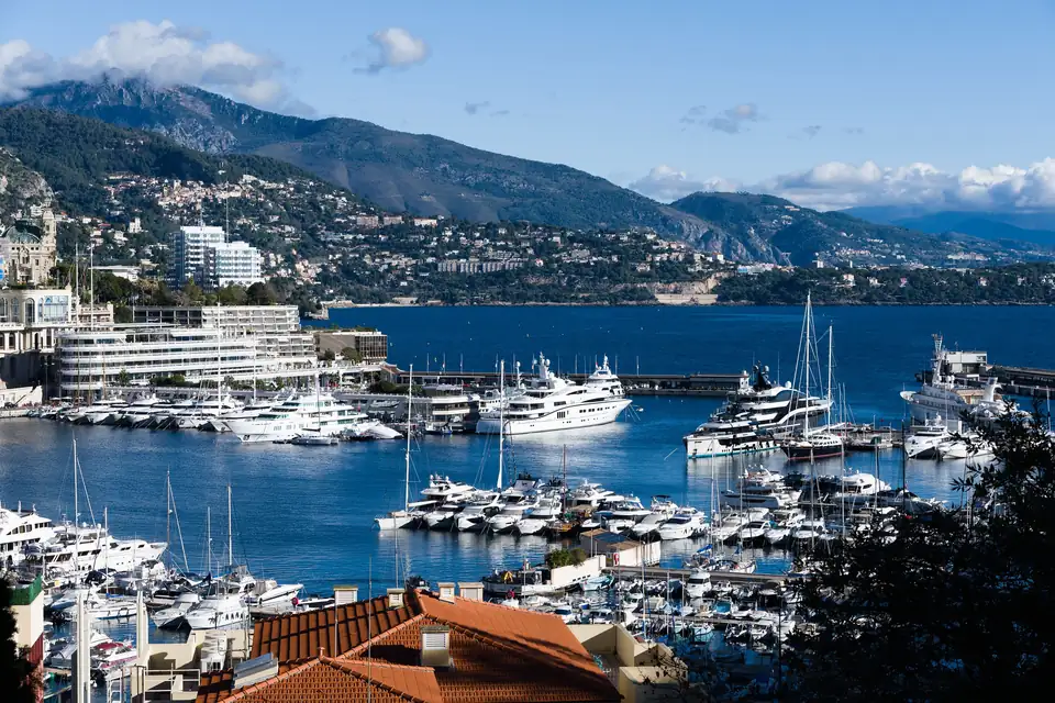 Superyachts and sailing yachts berthed in Monaco's Port Hercule, below terracotta rooftops, with the hills of the French Riviera beyond