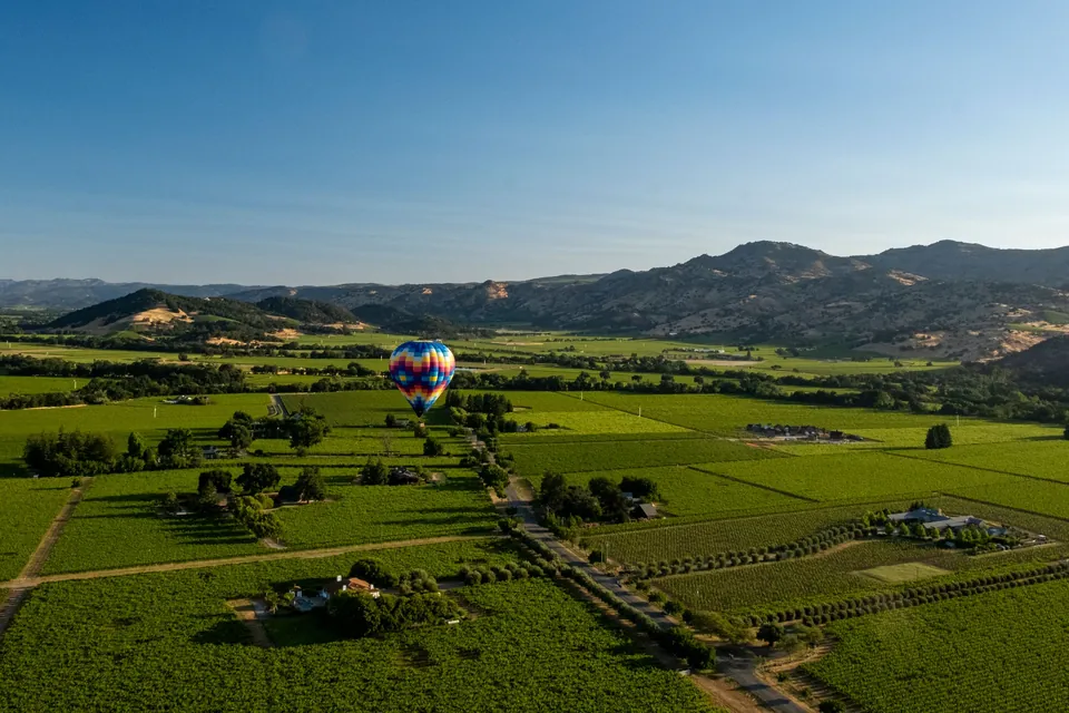 A rainbow-patterned hot-air balloon drifting low over green vineyards and wine estates in Napa Valley, California, with oak-dotted hills beyond
