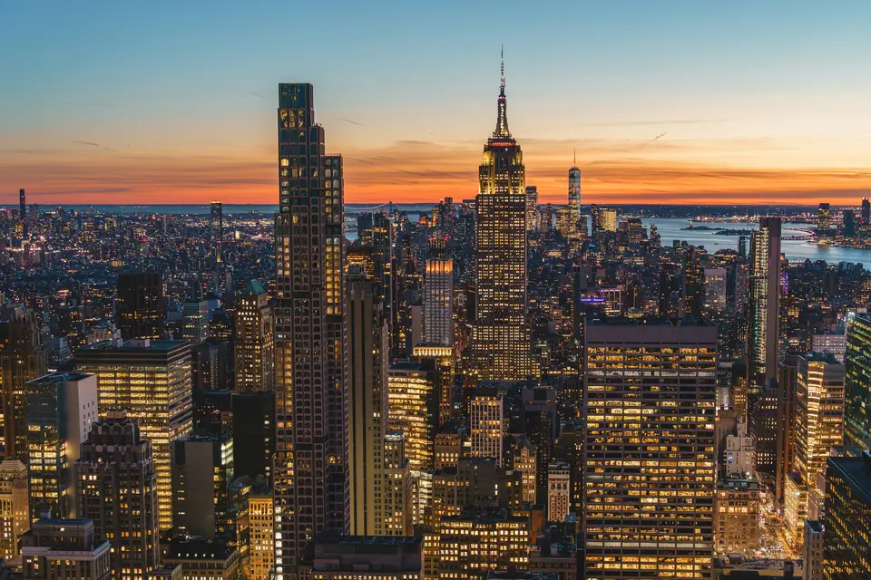 Midtown Manhattan from above at dusk, its towers lit gold, with the Empire State Building against an orange sky and the harbor in the distance
