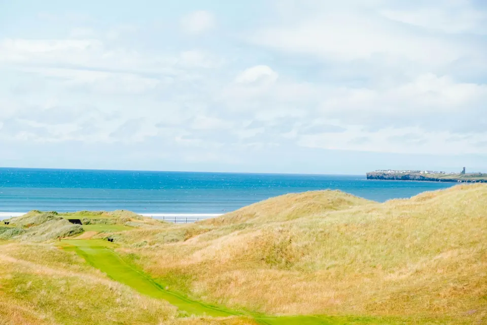 A links golf hole winding through golden dune grass above a blue bay on the Irish coast
