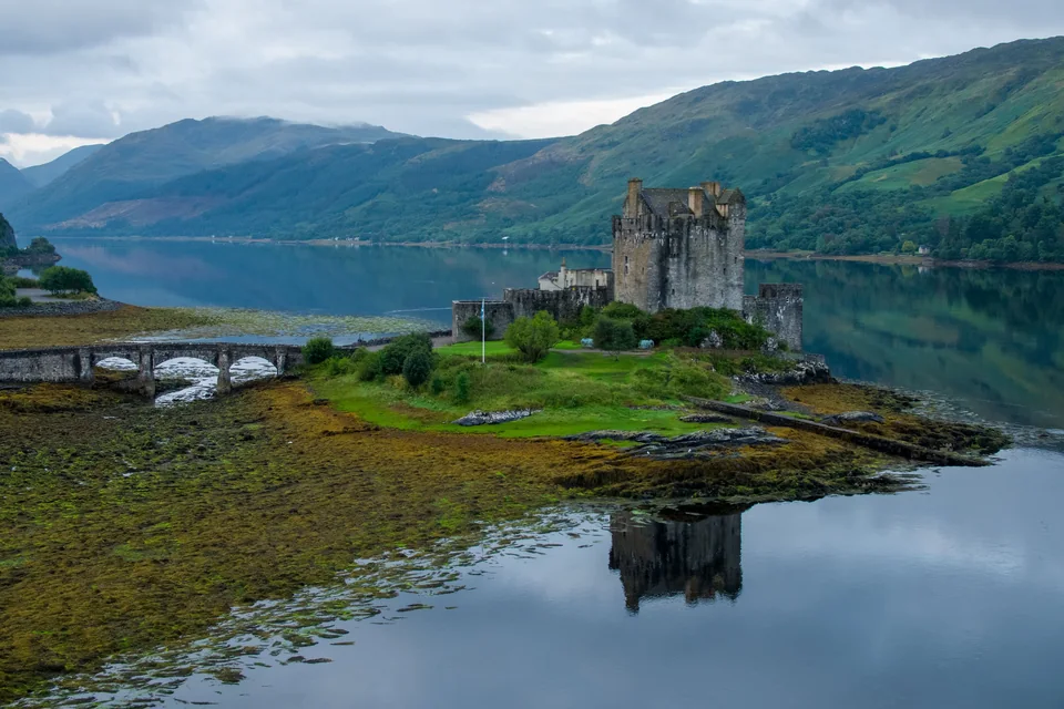 Eilean Donan Castle on its island in Loch Duich, reached by a stone footbridge and reflected in still water below green Highland hills, Scotland