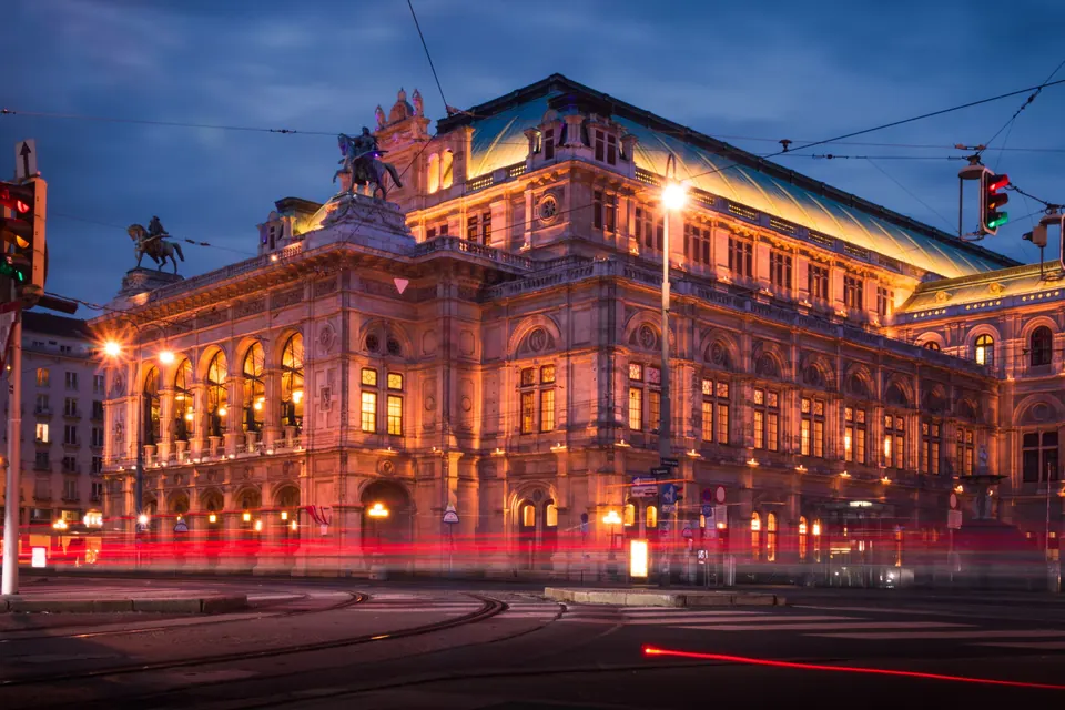The Vienna State Opera at blue hour, its facade lit warm gold above red light trails from passing traffic