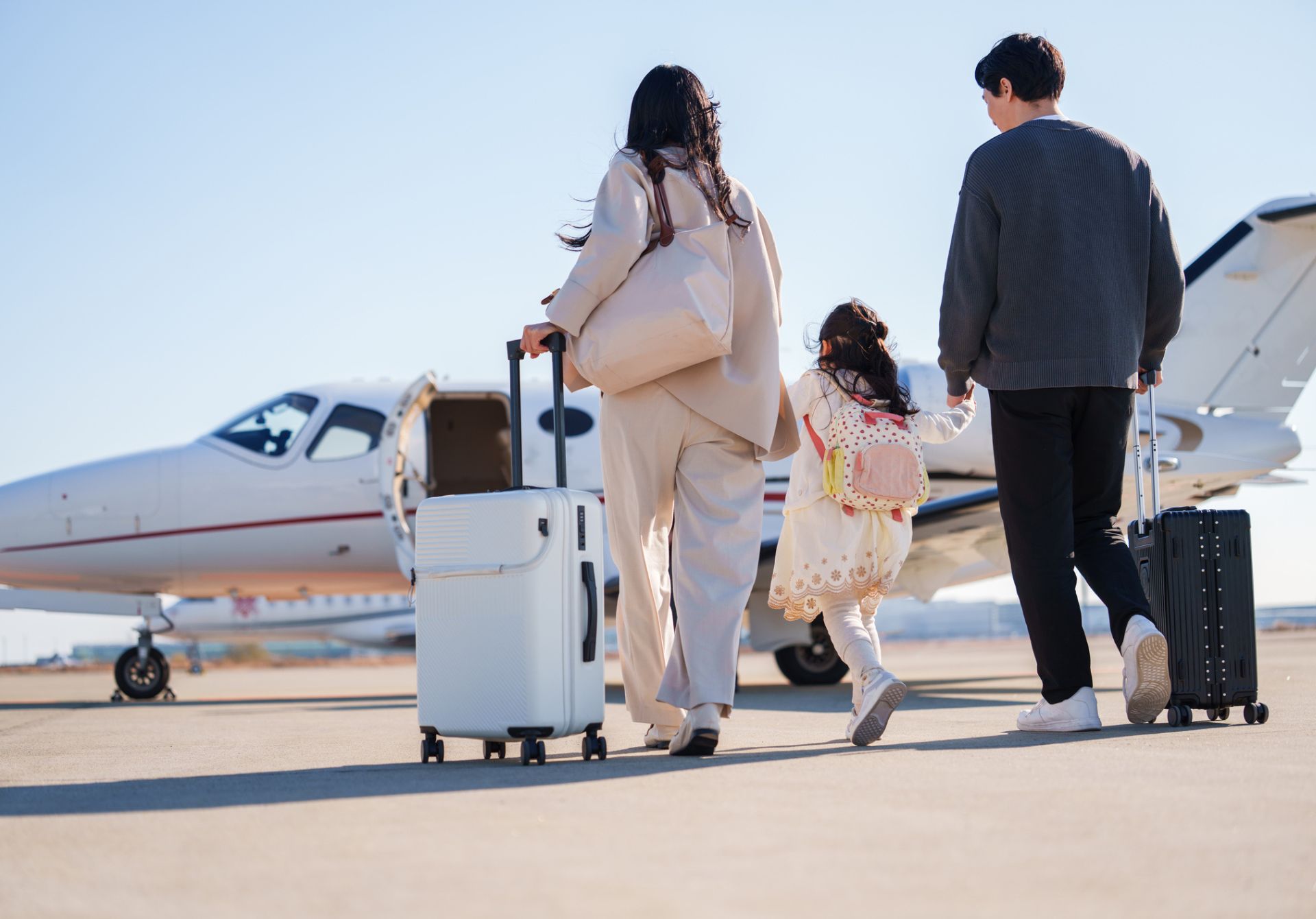 Family of three walking toward a small private jet with rolling suitcases on a tarmac under a clear blue sky.