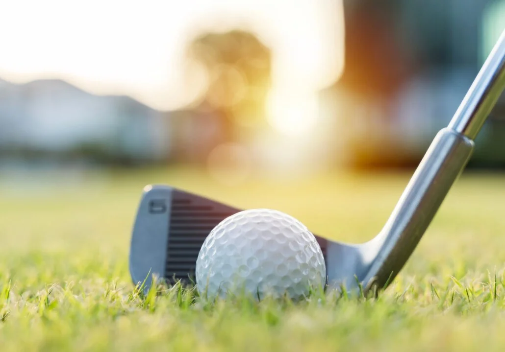 Golf ball on grass with a golf iron behind it and a warm, sunlit bokeh background.
