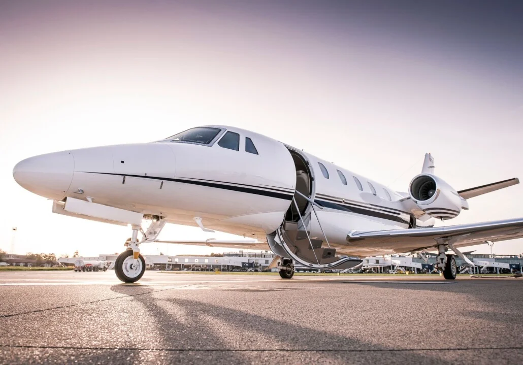 White private jet sits on the tarmac at sunset with the door open and boarding stairs visible.