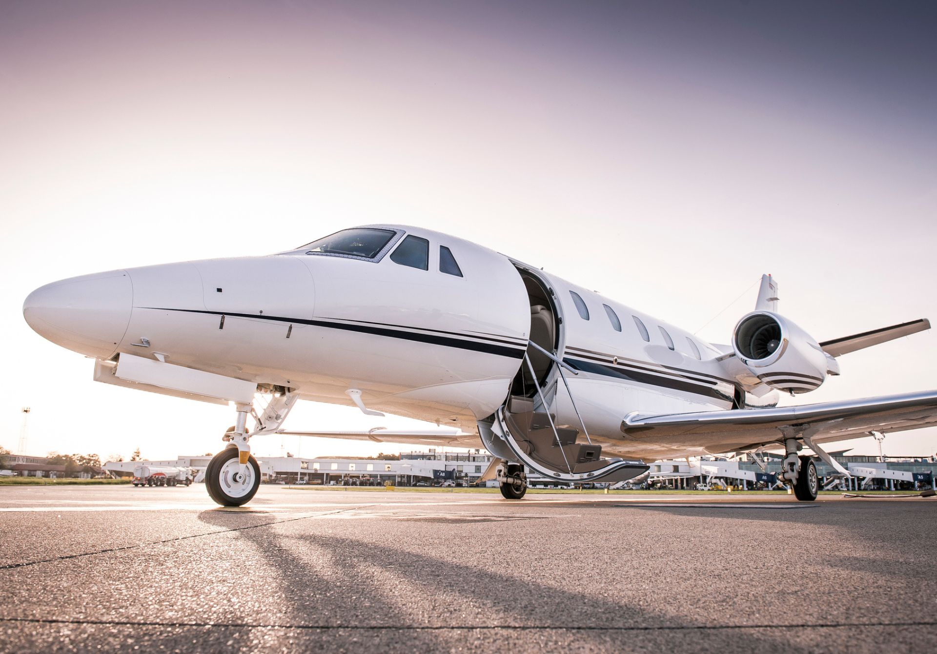 White private jet sits on the tarmac at sunset with the door open and boarding stairs visible.